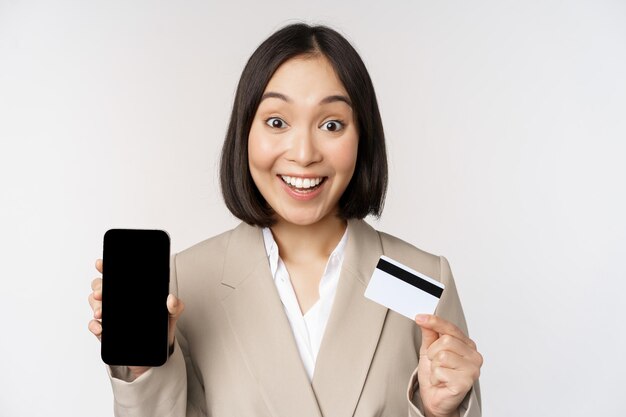 Geschäftsfrau mit fröhlichem, enthusiastischem Gesicht, das Kreditkarten- und Smartphone-App-Bildschirm zeigt, der im Anzug vor weißem Hintergrund steht