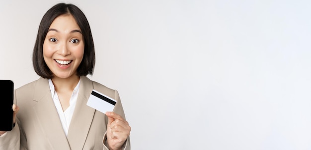 Geschäftsfrau mit fröhlichem, enthusiastischem Gesicht, das Kreditkarten- und Smartphone-App-Bildschirm zeigt, der im Anzug vor weißem Hintergrund steht