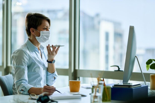 Geschäftsfrau mit Gesichtsmaske, die an einem Computer arbeitet, während sie Sprachnachrichten auf dem Smartphone im Büro aufzeichnet