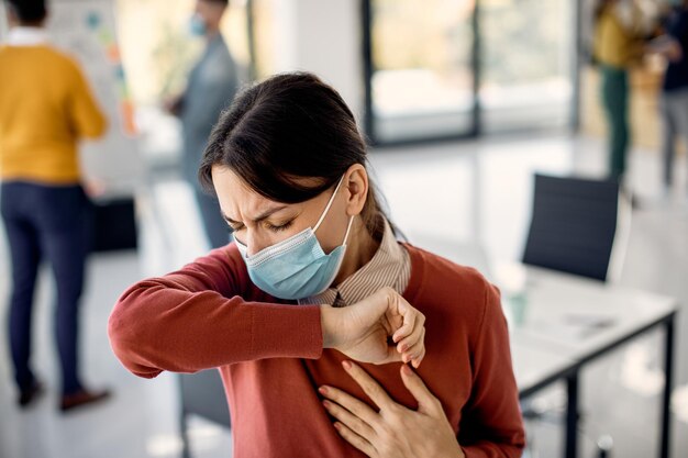 Geschäftsfrau mit Gesichtsmaske, die während der Arbeit im Büro in den Ellbogen hustet