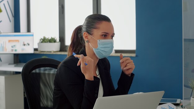 Geschäftsfrau mit medizinischer Gesichtsmaske, die mit ihrem Team über Kommunikationsstrategie am Schreibtisch spricht. Das Geschäftsteam hält die soziale Distanzierung aufrecht, während es in einem neuen normalen Firmenbüro arbeitet