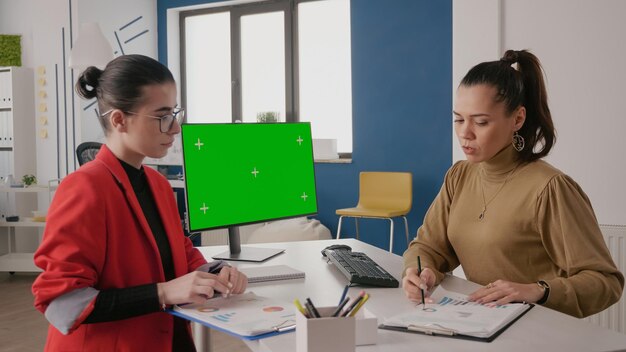 Geschäftsfrauen, die einen Computer mit grünem Bildschirm am Schreibtisch verwenden. Team von Leuten, die sich unterhalten, während sie mit Mock-up-Hintergrund und isolierter Vorlage auf dem Computerbildschirm arbeiten. Chroma-Key-Kopierbereich