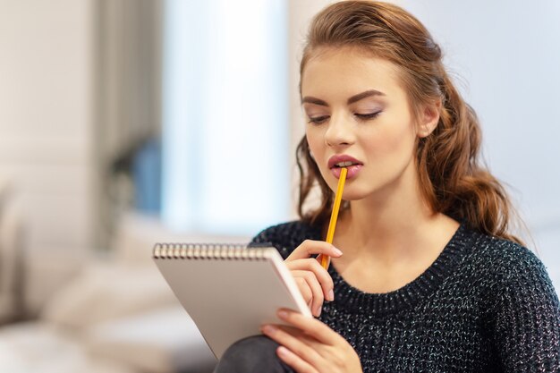 Geschäftsideen. Zu Hause studieren und arbeiten. Nachdenkliche junge Frau, die Notizen mit Notizblock in der Küche macht.