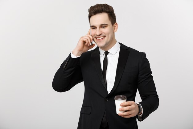 Geschäftskonzept: Nahaufnahme überzeugter junger gutaussehender Geschäftsmann, der auf Handy spricht und Kaffee über weißem lokalisiertem Hintergrund trinkt.