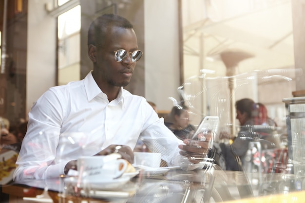 Geschäftsleute, moderner urbaner Lebensstil und Technologien. Hübscher selbstbewusster afroamerikanischer Geschäftsmann in den Schattierungen und im weißen Hemd, die SMS schreiben oder E-Mail auf Handy während der Kaffeepause im Café abrufen