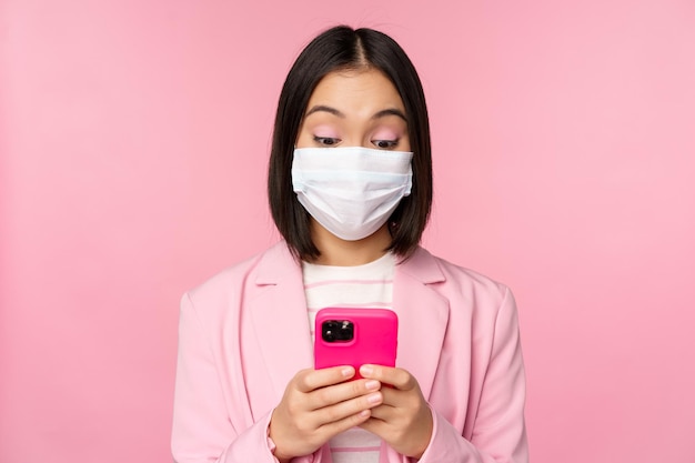 Geschäftsleute und Covid19-Konzept Junge asiatische Geschäftsfrau in Anzug und medizinischer Gesichtsmaske mit Smartphone-App, die über rosafarbenem Hintergrund steht