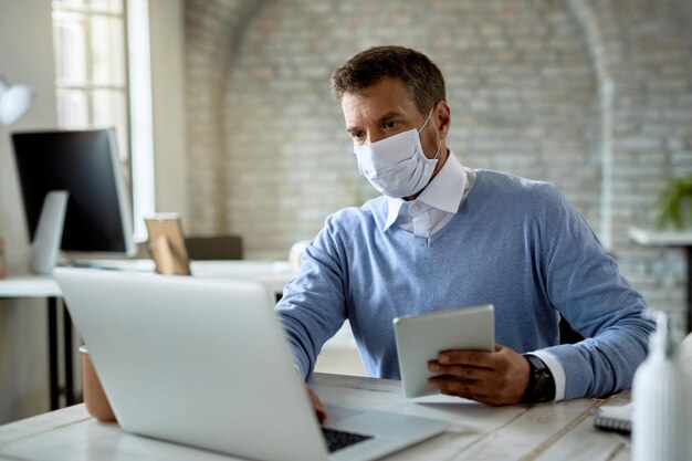 Geschäftsmann, der eine schützende Gesichtsmaske trägt, während er an einem Computer arbeitet und ein digitales Tablet im Büro verwendet