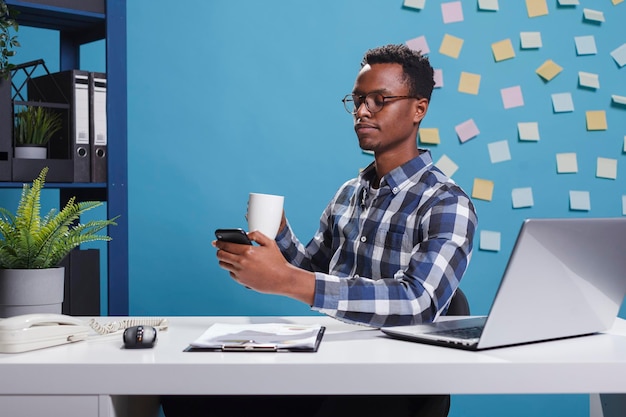 Geschäftsmann der Marketingfirma, der soziale Medien auf dem mobilen Smartphone durchsucht, während er eine Tasse Kaffee trinkt. Start-up-Projektmanager benachrichtigt Leute über Mobiltelefon, während er eine Tasse Tee genießt.