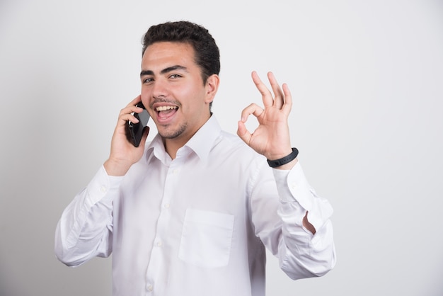 Geschäftsmann, der mit Handy spricht und ok Zeichen auf weißem Hintergrund zeigt.