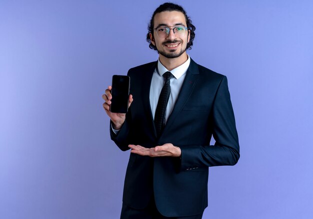 Geschäftsmann im schwarzen Anzug und in der Brille, die das Smartphone zeigt, das es mit Arm seiner Hand präsentiert, der zuversichtlich über der blauen Wand steht