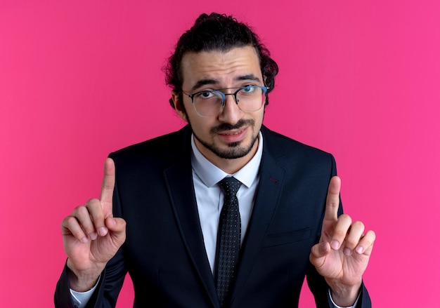 Geschäftsmann im schwarzen Anzug und in der Brille, die nach vorne schauen und Zeigefinger zeigen, die über rosa Wand stehen