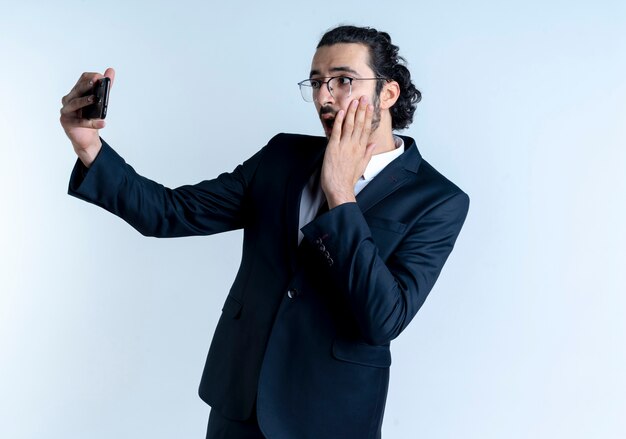 Geschäftsmann im schwarzen Anzug und in der Brille, die selfie unter Verwendung seines Handys nehmen, das überrascht steht, über weißer Wand stehend