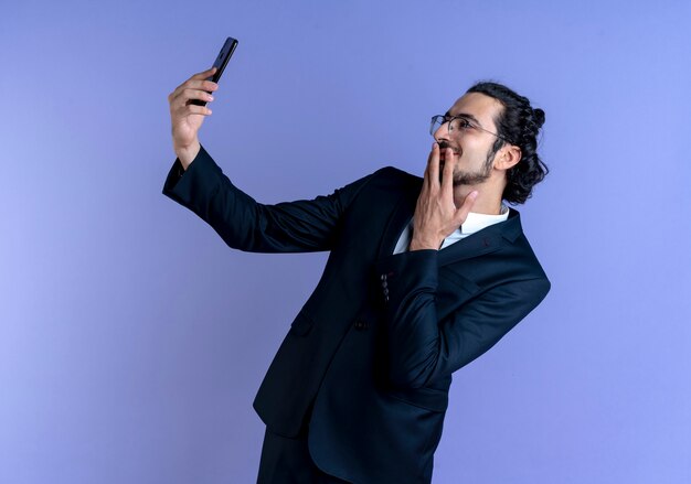 Geschäftsmann im schwarzen Anzug und in der Brille, die selfie unter Verwendung seines Smartphones nehmen, das nach vorne mit schüchternem Lächeln auf Gesicht steht, das über blauer Wand steht