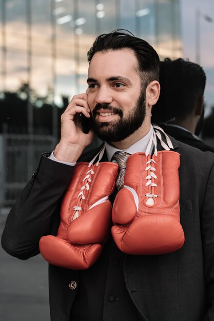 Geschäftsmann in einer Klage sprechend an seinem Handy mit den roten Boxhandschuhen, die von seinem Halslehnen hängen
