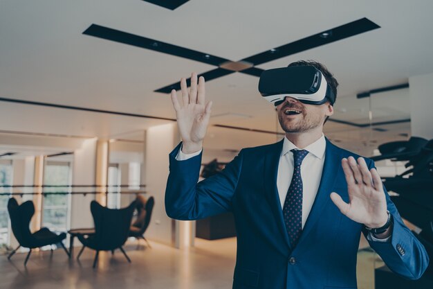 Geschäftsmann in futuristischem vr-headset berührt die luft mit seinen handflächen, die in die virtuelle realität eingetaucht sind. junger finanzier mit innovativer methode des spielerlebnisses in der bürolobby während der kaffeepause