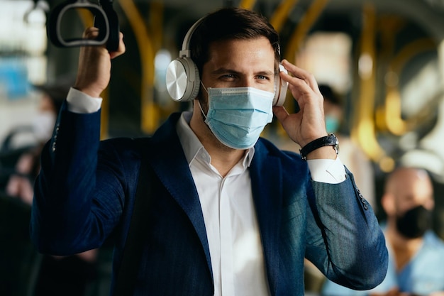 Geschäftsmann mit Gesichtsmaske, der Musik über Kopfhörer hört, während er mit dem Bus zur Arbeit fährt
