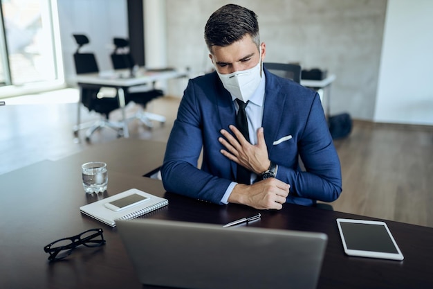 Geschäftsmann mit Gesichtsmaske, der Schmerzen in der Brust verspürt, während er im Büro am Laptop arbeitet