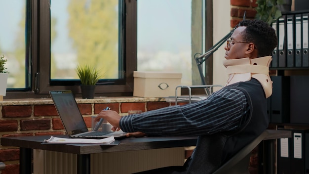 Geschäftsmann mit Halskragen bei der Arbeit nach Wirbelverletzung im Startup-Büro. Verletzter Mitarbeiter, der medizinischen Schaum verwendet, während er mit einem Laptop arbeitet, um kommerzielle Investitionen zu planen.