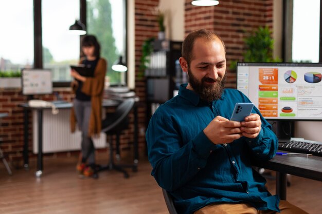 Geschäftsmann mit Smartphone, der auf Social Media-SMS mit entfernten Freunden im Büro des Startup-Unternehmens schaut. Manager sucht nach Geschäftsinformationen und entwickelt eine Online-Kommunikations-App.