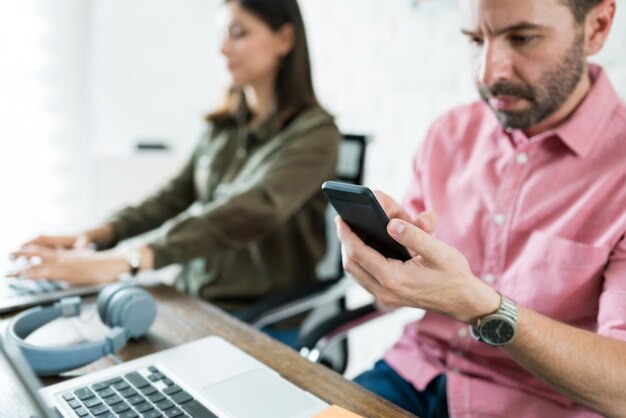 Geschäftsmann mittleren Alters schreibt SMS auf Smartphone, während er mit Kollege am Schreibtisch im Büro sitzt