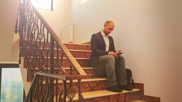 Geschäftsmann sitzt allein auf der Treppe und tippt auf dem Smartphone in der Finanzgesellschaft, die Überstunden macht. Gruppe professioneller erfolgreicher Geschäftsleute, die im modernen Finanzgebäude arbeiten.