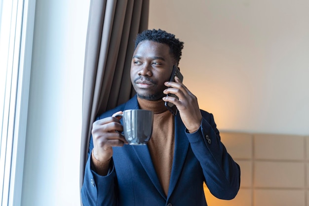 Geschäftsmann stand am Fenster im Hotelzimmer und trank Kaffee Nachdenklicher junger afrikanischer Geschäftsmann stand am Fenster mit einer Tasse Kaffee und schaute auf sein Handy