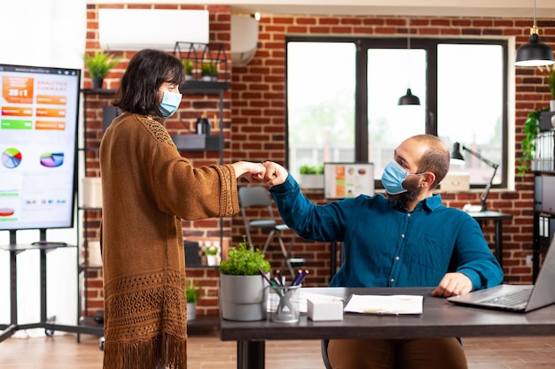 Geschäftsteam mit medizinischer Gesichtsschutzmaske gegen Coronavirus-Gruß, der den ersten Stoß im Startup-Büro gibt. Manager-Mann, der am Schreibtisch sitzt und Ideen für die Tippstrategie von Geschäftsprojekten sammelt