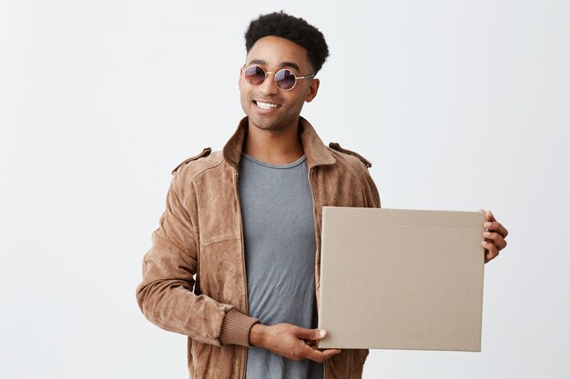 Geschenk für Sie. Isoliert auf weißem Porträt des jungen modischen dunkelhäutigen Mannes mit Afro-Frisur in grauem T-Shirt, brauner Jacke und Sonnenbrille, die Box in der Hand hält und in der Kamera lächelt.