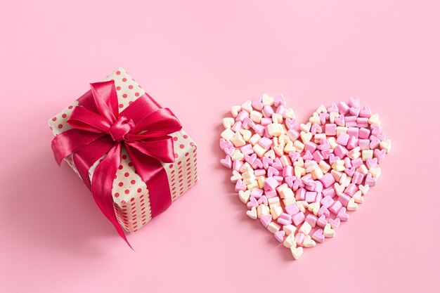 Geschenkbox mit roter Schleife und Herz aus Bonbons auf rosa Oberfläche