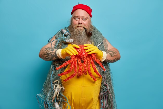 Geschickter bärtiger Fischer hält großen Tintenfisch in Händen, die mit Fischernetz gefangen werden, posiert im Hafen, trägt gelbe Gummihandschuhe