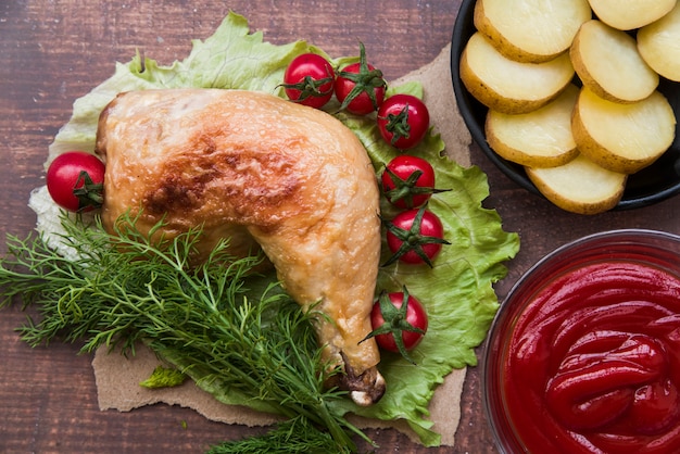 Kostenloses Foto geschnittene gekochte kartoffeln; gebratene hühnerkeule; soße; kirschtomate; dill zum abendessen auf salatblatt über holztisch