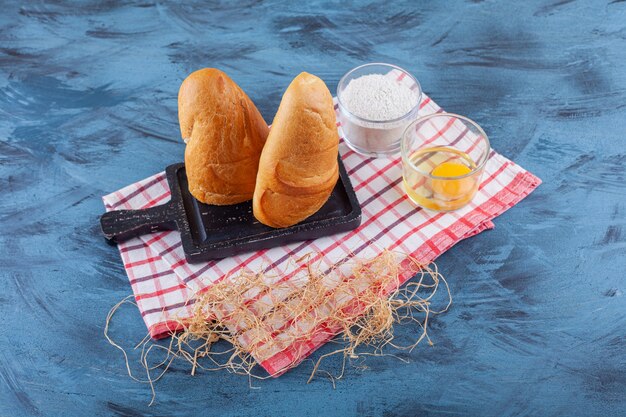 Geschnittenes Brot auf einem Brett neben Mehl und Ei auf einem Geschirrtuch auf der blauen Oberfläche.