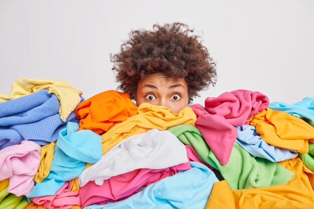 Geschockte Frau mit lockigem Afro-Haar starrt verwanzte Augen an, die in einem riesigen Haufen bunter Kleidung ertrunken sind, räumt Schrank aus wählt Kleidung für Spenden oder Recycling-Weiß aus