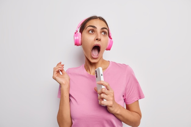 Geschockter weiblicher meloman hält den mund weit geöffnet und konzentriert sich oben mit wunder hält handy hört musik in drahtlosen stereokopfhörern trägt lässiges rosa t-shirt isoliert über weißer wand