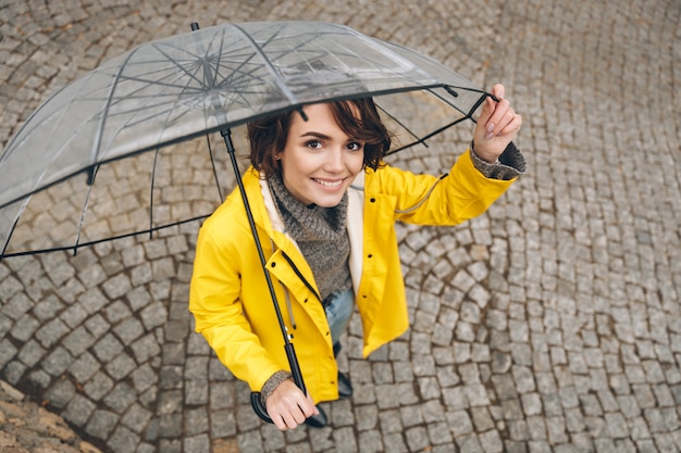 Geschossen von oben genanntem der wunderbaren Frau im gelben Regenmantel, der beim Gehen unter großen transparenten Regenschirm glücklich ist