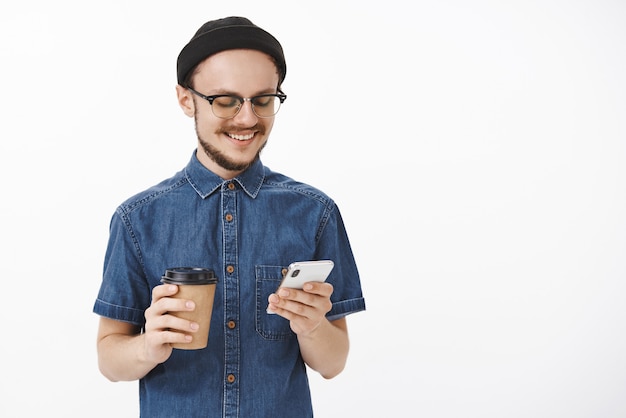 geselliger glücklicher und entspannter junger stilvoller moderner und unrasierter Kerl in der schwarzen Mütze der Gläser und im blauen Hemd, die Smartphone und Pappbecher Kaffee halten, der freudig auf Gerätebildschirm lächelt