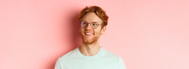 Kostenloses Foto gesicht eines gutaussehenden rothaarigen mannes mit brille, der erfreut lächelt und die obere rechte ecke betrachtet, die über ihm steht