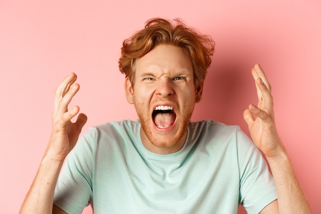 Kostenloses Foto gesicht eines wütenden rothaarigen mannes, der wütend schreit und die hände schüttelt, empört und fluchend anstarrt, hass und aggression ausdrückt und frustriert über rosa hintergrund steht