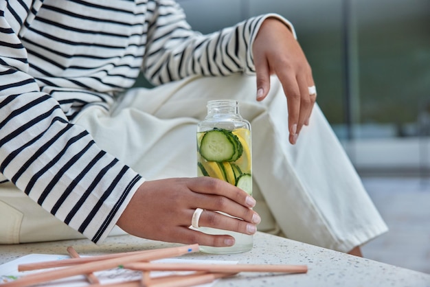 Gesichtslose Frau in Freizeitkleidung hält eine Flasche Detox-Trinkwasser mit Zitronenscheiben und Gurkenresten nach dem Zeichnen eines von Buntstiften umgebenen Bildes