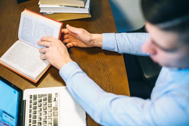 Gesichtsloser Mann, der Buch bei der Anwendung des Laptops nimmt