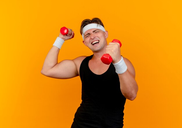 Gespannter junger hübscher sportlicher Mann, der Stirnband und Armbänder trägt, die Hanteln an der orange Wand lokalisiert halten