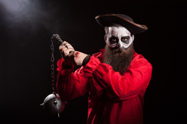 Gespenstischer männlicher Pirat mit dem langen Bart, der eine Keule über schwarzem Hintergrund hält. Halloween-Outfit.