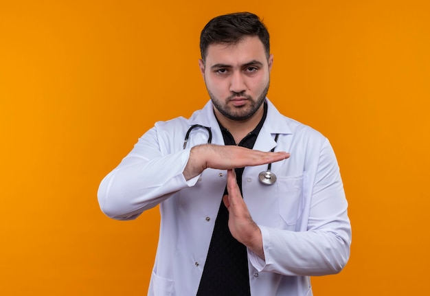 Gestörter junger bärtiger männlicher Arzt, der weißen Kittel mit Stethoskop trägt und Kamera unzufrieden betrachtet, die Auszeitgeste mit Händen macht