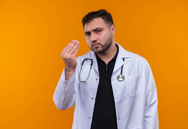 Gestörter junger bärtiger männlicher Arzt, der weißen Kittel mit Stethoskop trägt und unzufrieden mit der Hand gestikuliert