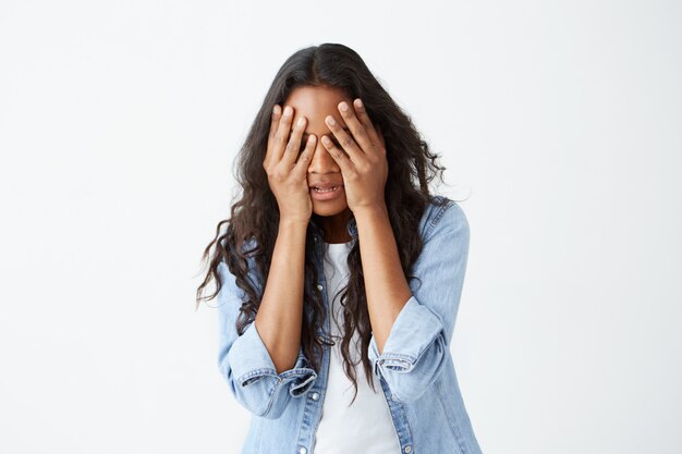 Gestresste afroamerikanische Frau mit langen, welligen Haaren, lässig gekleidet, verärgert, die Hände auf dem Kopf haltend und ihr Gesicht verzweifelt versteckt, nachdem sie schlechte Nachrichten gehört hatte.