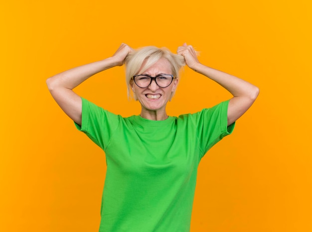 Gestresste blonde slawische Frau mittleren Alters, die eine Brille trägt, die Front zieht, die Haare auf gelber Wand isoliert zieht