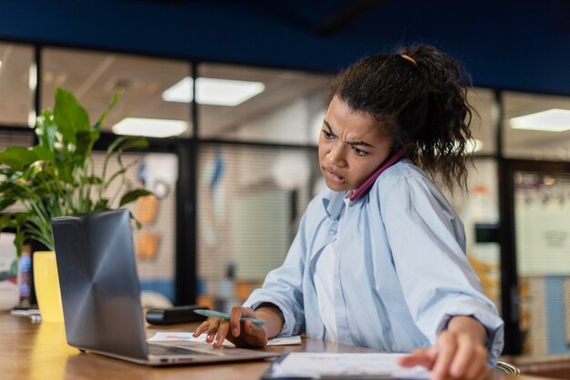 Gestresste Frau im Büro mit Laptop und Gespräch auf dem Smartphone