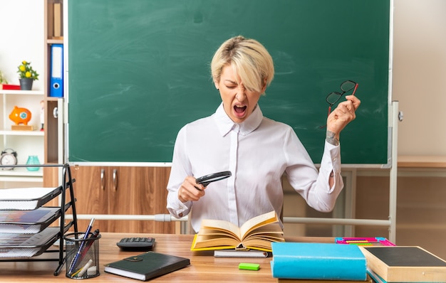 Gestresste junge blonde Lehrerin, die am Schreibtisch mit Schulwerkzeugen im Klassenzimmer sitzt und eine Lupe über dem offenen Buch hält, die Brille abnimmt und mit geschlossenen Augen schreit