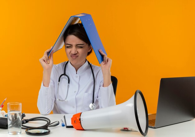 Gestresste junge Ärztin, die medizinische Robe und Stethoskop trägt, sitzt am Schreibtisch mit medizinischem Werkzeuglautsprecher und Laptop, die Ordner auf Kopf mit geschlossenen Augen isoliert auf gelber Wand halten