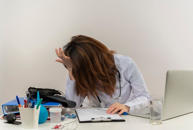 Gestresste Ärztin mittleren Alters, die medizinische Robe und Stethoskop trägt, sitzt am Schreibtisch mit Zwischenablage des medizinischen Werkzeugs und Laptop, die Hände auf Kopf und auf Schreibtisch isoliert setzen